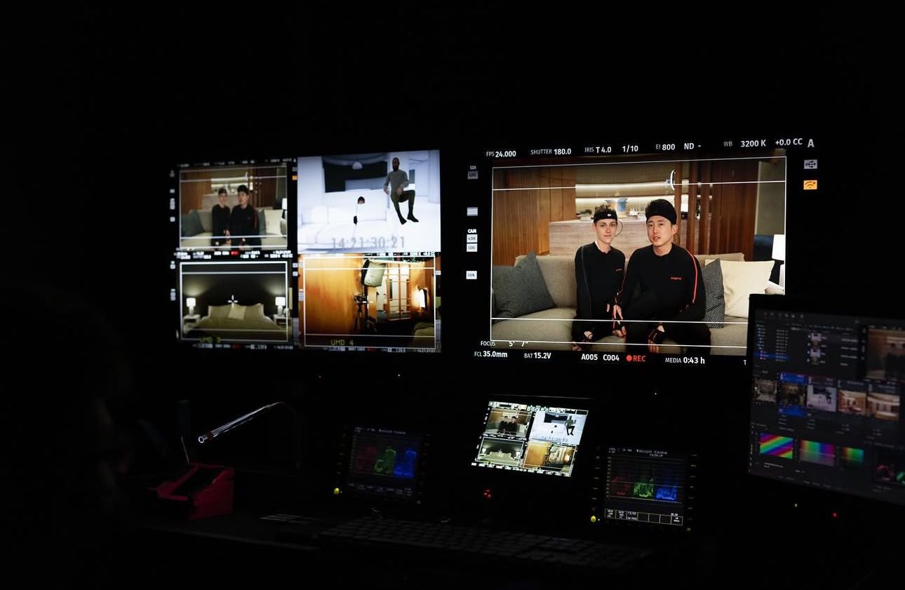 Two persons on the Love Me set are sitting on a couch displayed on a monitor for BTS